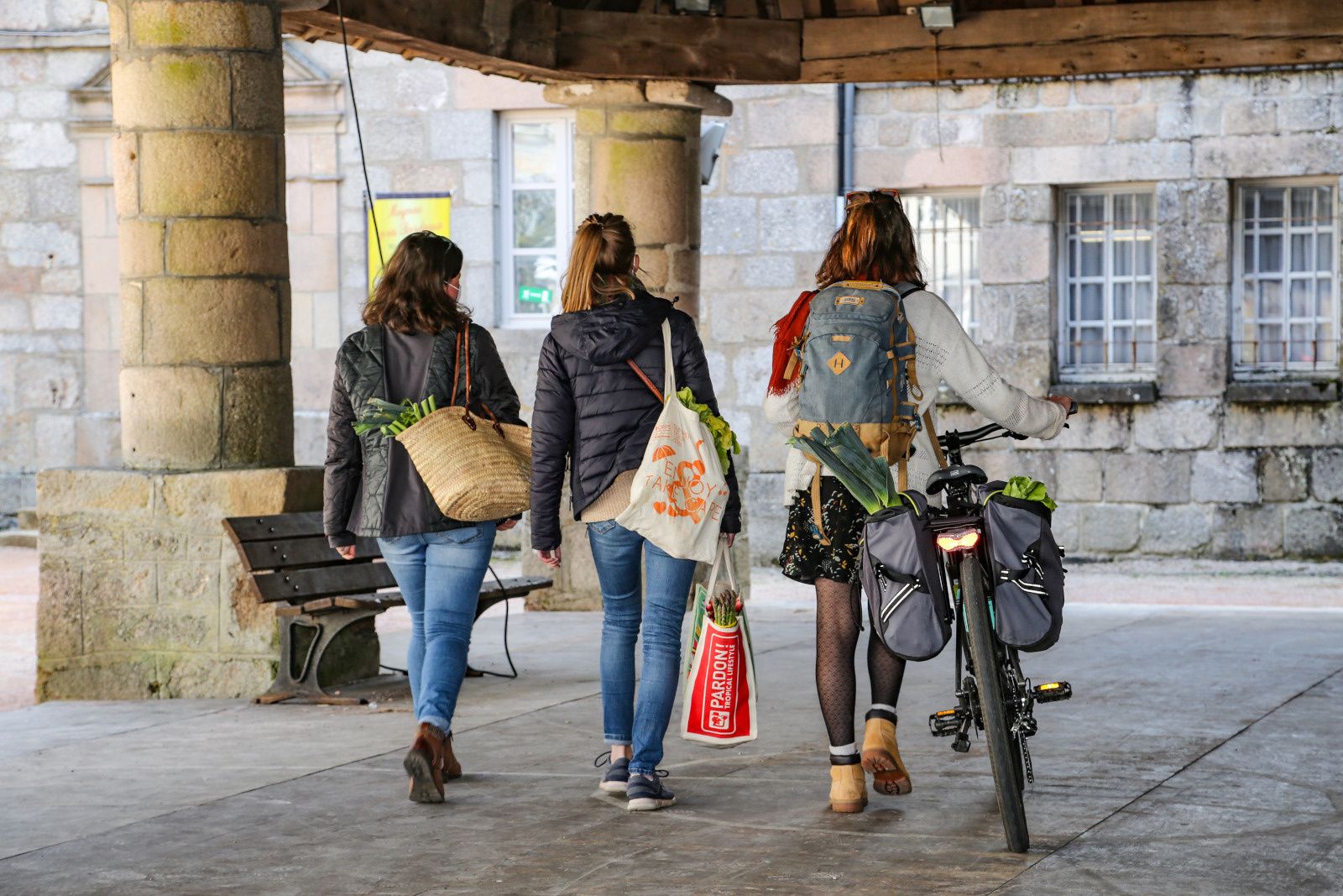 3 personnes de dos avec des sacs en tissus et en paille pleins de fruits et légumes. 2 personnes sont à pied et la 3ème marche à côté en tenant son vélo